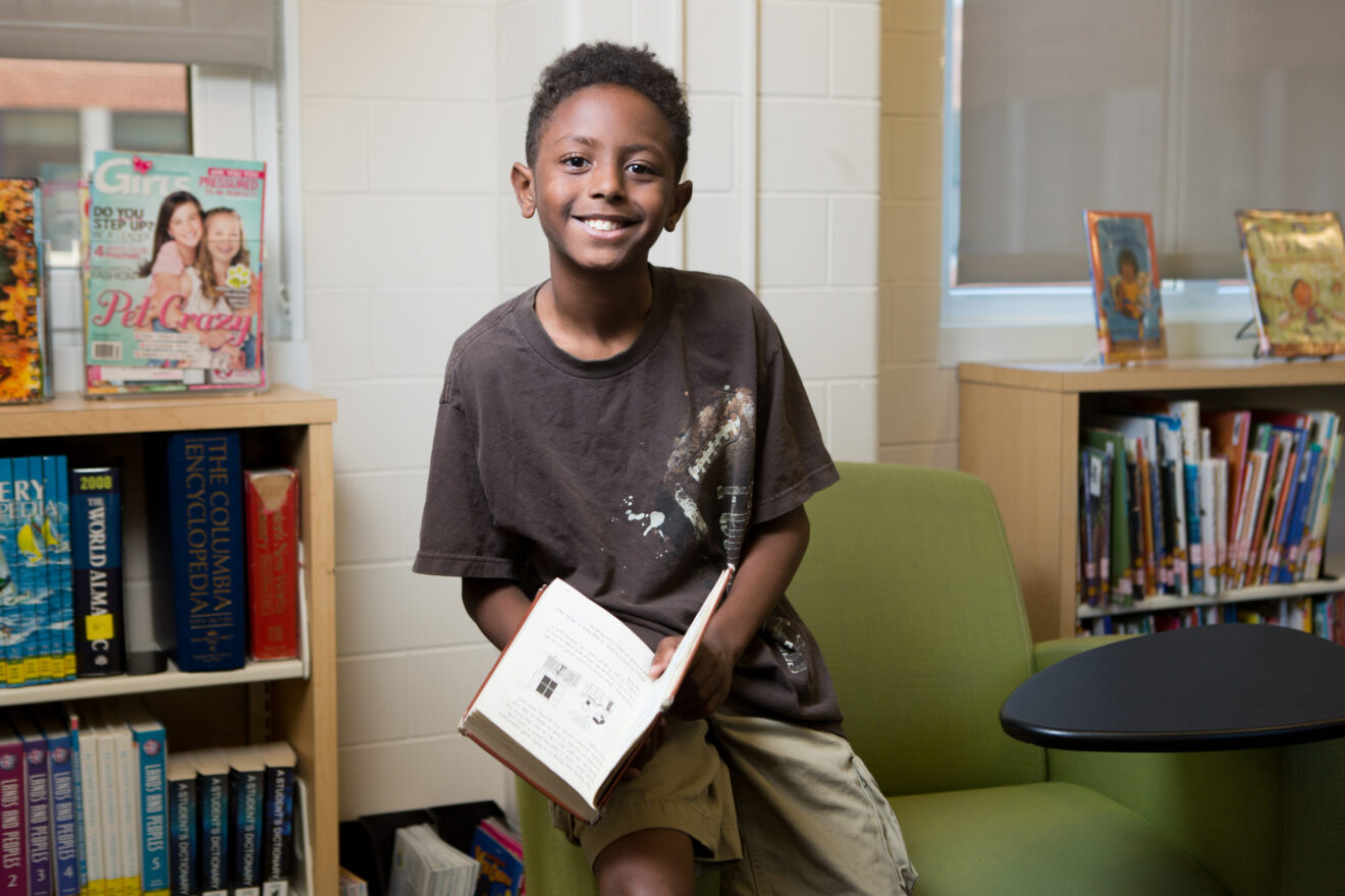 Black male middle-school student with book sitting on arm of chair and smiling for literacy program article
