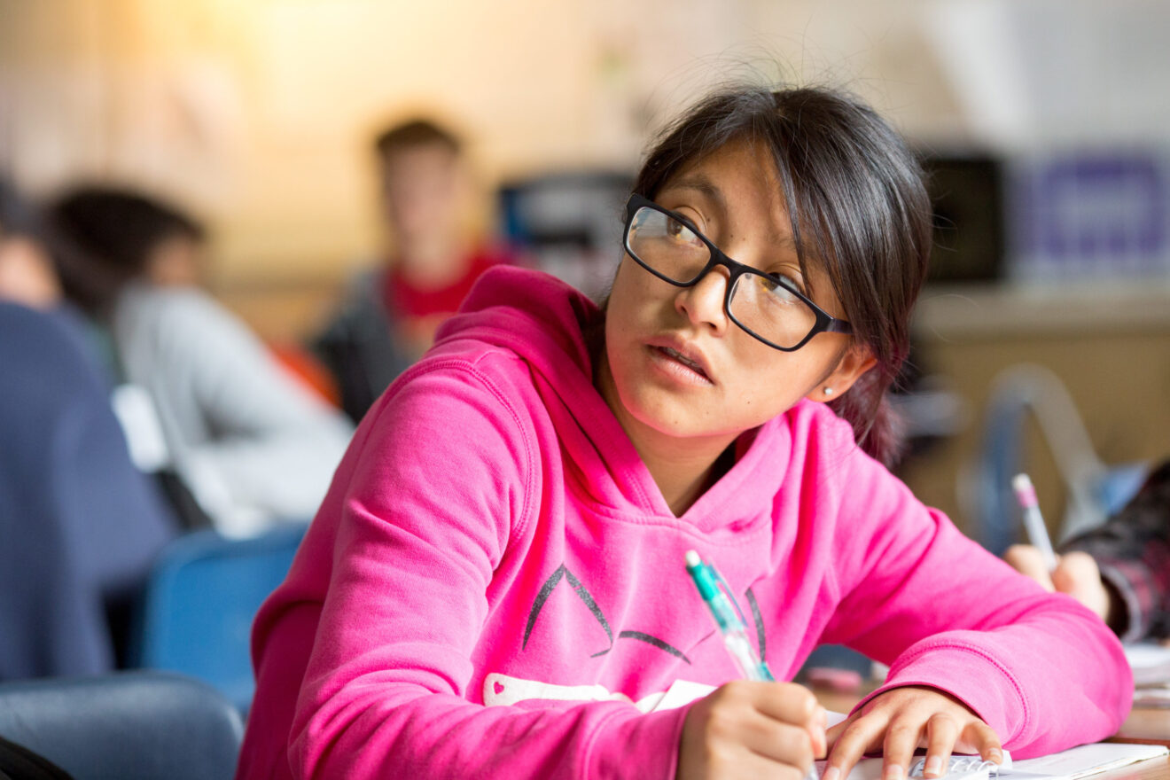 Hispanic girl writing in a notebook looking over her shoulder in the classroom - for bilingual education article.