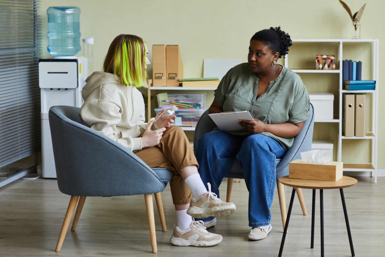 Black female social worker talking to teenage girl while they sitting on armchairs at office for article on mental health supports