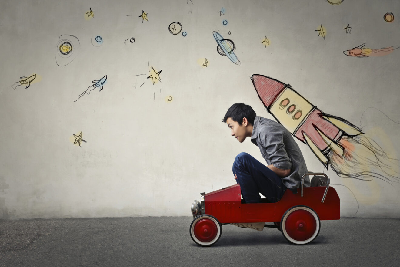 Young Asian male squished into a red toy car in front of a gray wall with spaceship and planets, for article on student momentum