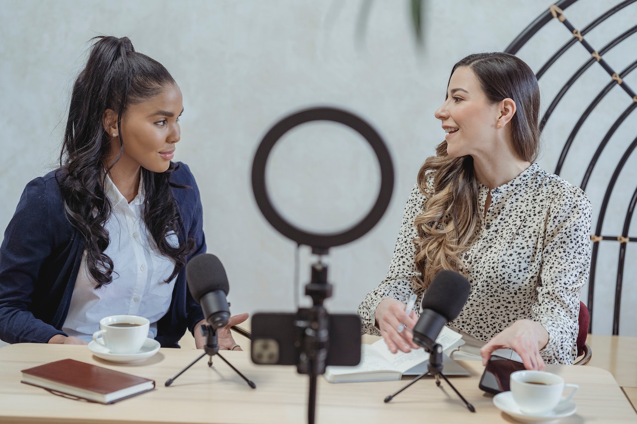 Two women recording a podcast and drinking coffee in front of a ring light