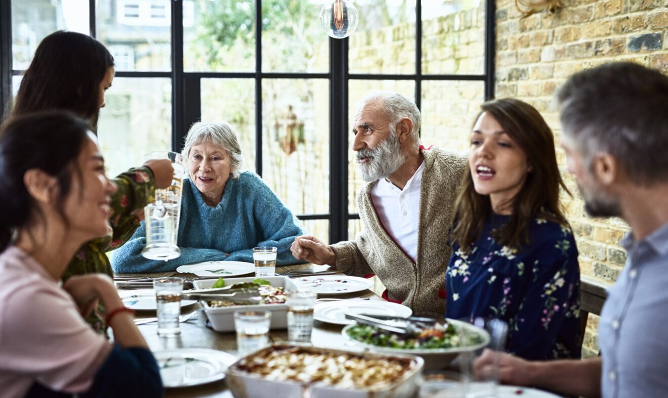 Mixed age ranges enjoying meal together in family home, pre teen girl pouring water for dinner guests, social vibrant lively family gathering