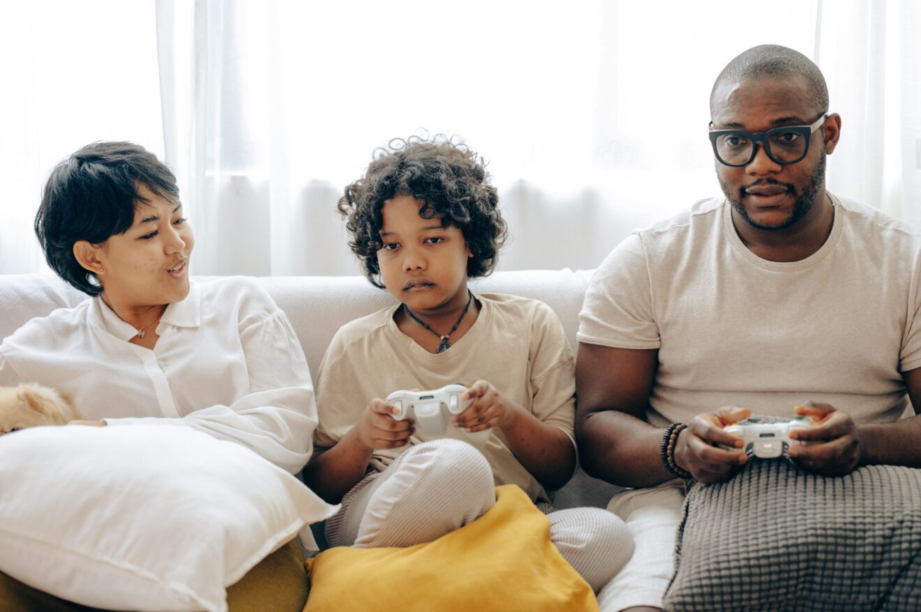 Black man and two kids playing video games while sitting on a couch.