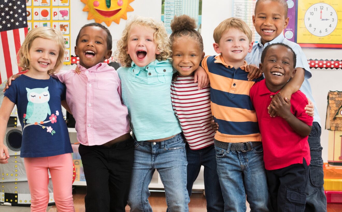 seven smiling preschoolers standing in a row in a classroom with arms around each other's shoulders.