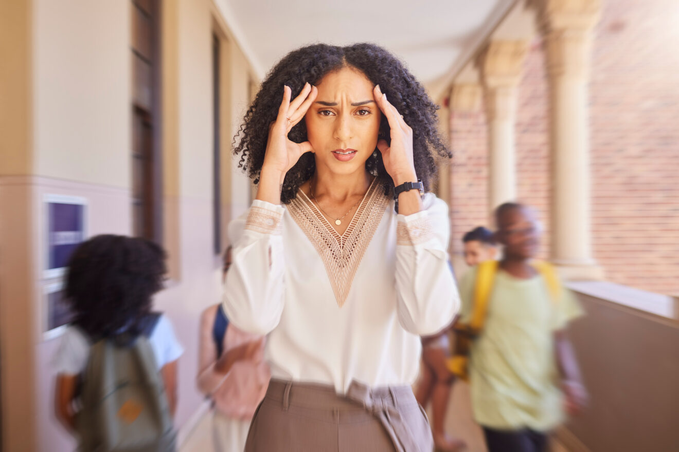 Black female teacher holding hands to her head as if she has a headache in the hallways of a school for article on what teachers need