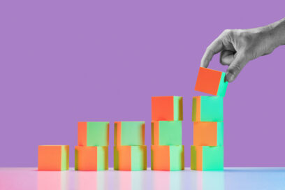 Man's hand in black and white placing block on largest stack of 5 growing stacks of yellow and red tinted blocks for article on teaching computer science