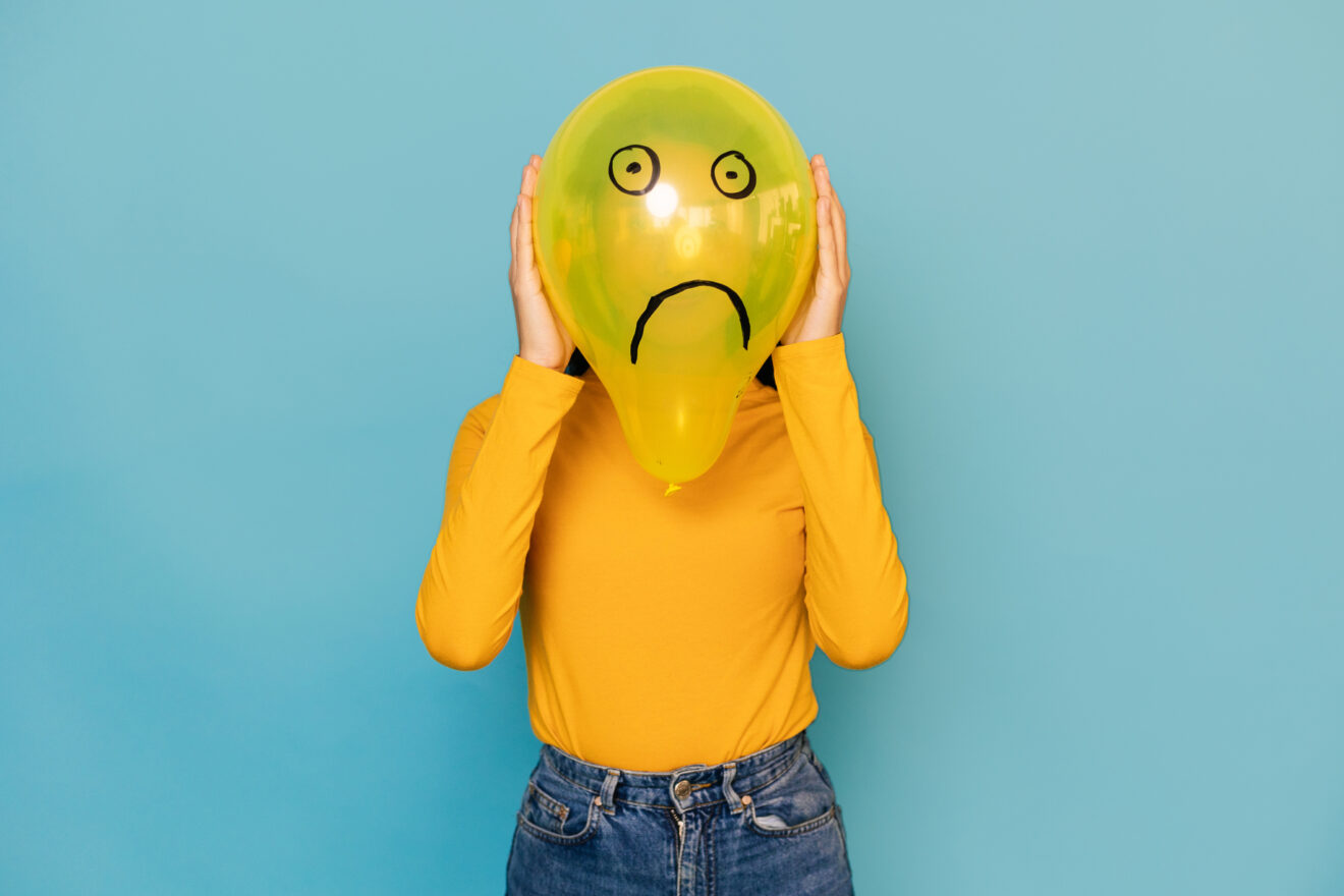 Young woman hidden behing a balloon with a sad face drawn on it over blue background. Negative emotion concept for article on mental health