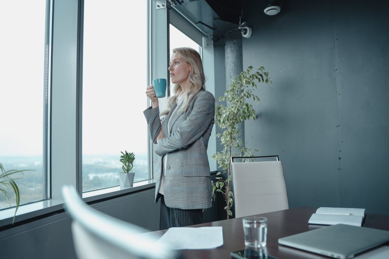 Boathouse Group’s 2024 survey on CEO attitudes on CMOs and marketing reveals four key findings. Pictured: A woman in a suit jacket standing, holding a cup of coffee in an office, looking out a window