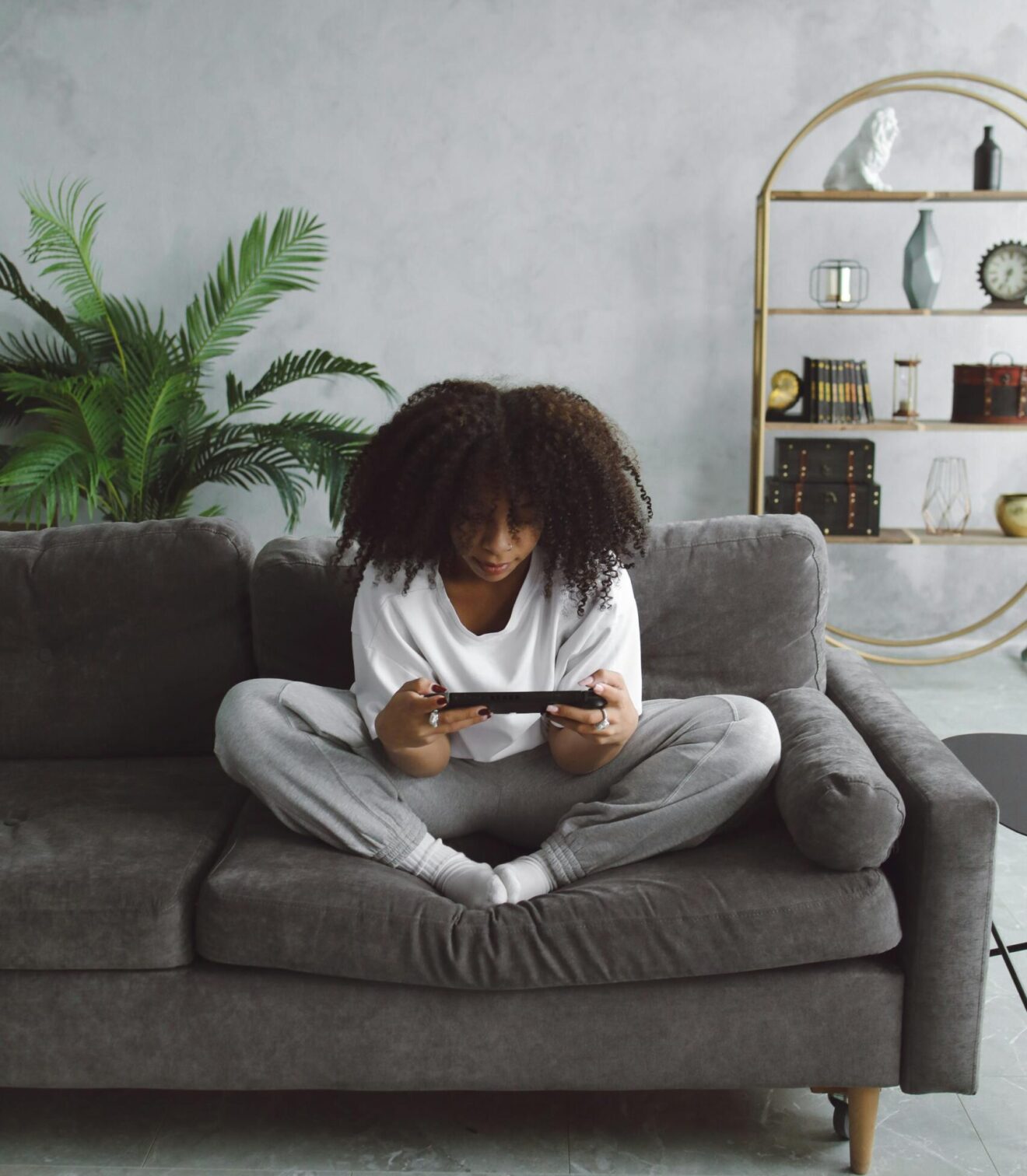 A young Black woman sits on a couch playing a handheld video game.