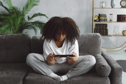 A young Black woman sits on a couch playing a handheld video game.