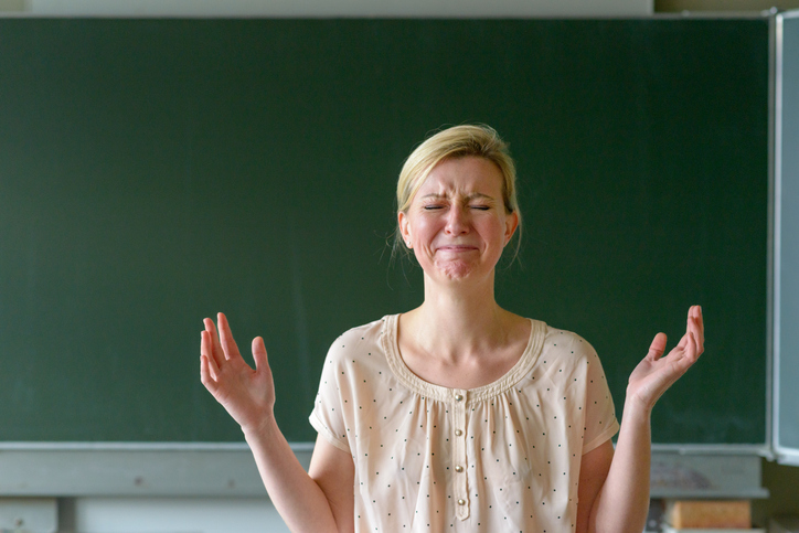 Angry frustrated school teacher with her hands up standing in front of a chalkboard in a classroom for article on teacher burnout