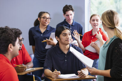 A multiracial group of six high school students sitting together in a classroom having a discussion, with a teacher leading. The students are in two teams, wearing either blue or red shirts. They could be participating in an after school activity, perhaps a debate team or math club. for article on career readiness