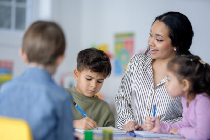 An Asian teacher encourages two young students as they draw and write at a sunny classroom table. The scene conveys learning, collaboration, and supportive, family-like education.