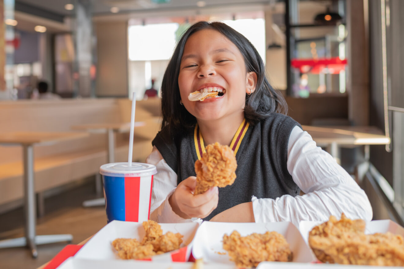Close up portrait of a satisfied little asian girl eating fried chicken and french fries In the restaurant. Unhealthy food concept, close up