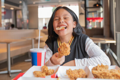 Close up portrait of a satisfied little asian girl eating fried chicken and french fries In the restaurant. Unhealthy food concept, close up