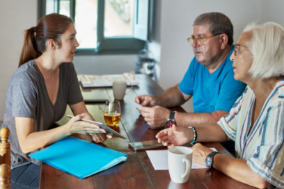 A young woman sitting at a wooden table across from an older couple, holding a tablet and gesturing during a conversation.