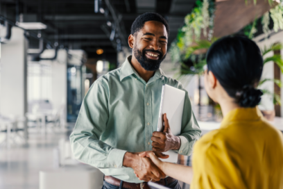 Smiling man holding papers and shaking hands with female in front of him.
