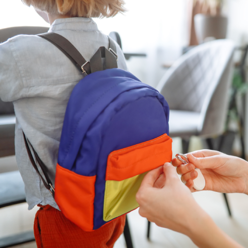 A close-up of a child wearing a colorful blue, orange, and yellow backpack while an adult's hands attach a white tracking tag on a carabiner to it.
