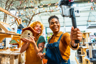 Two young women, shop assistants, are holding a birdhouse and smiling at camera during livestreaming in a pet shop Used for SmartBrief Marketing Original on small business content creation and AI