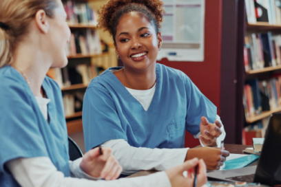 Two students in blue medical scrubs studying together at a library table with books and a laptop.