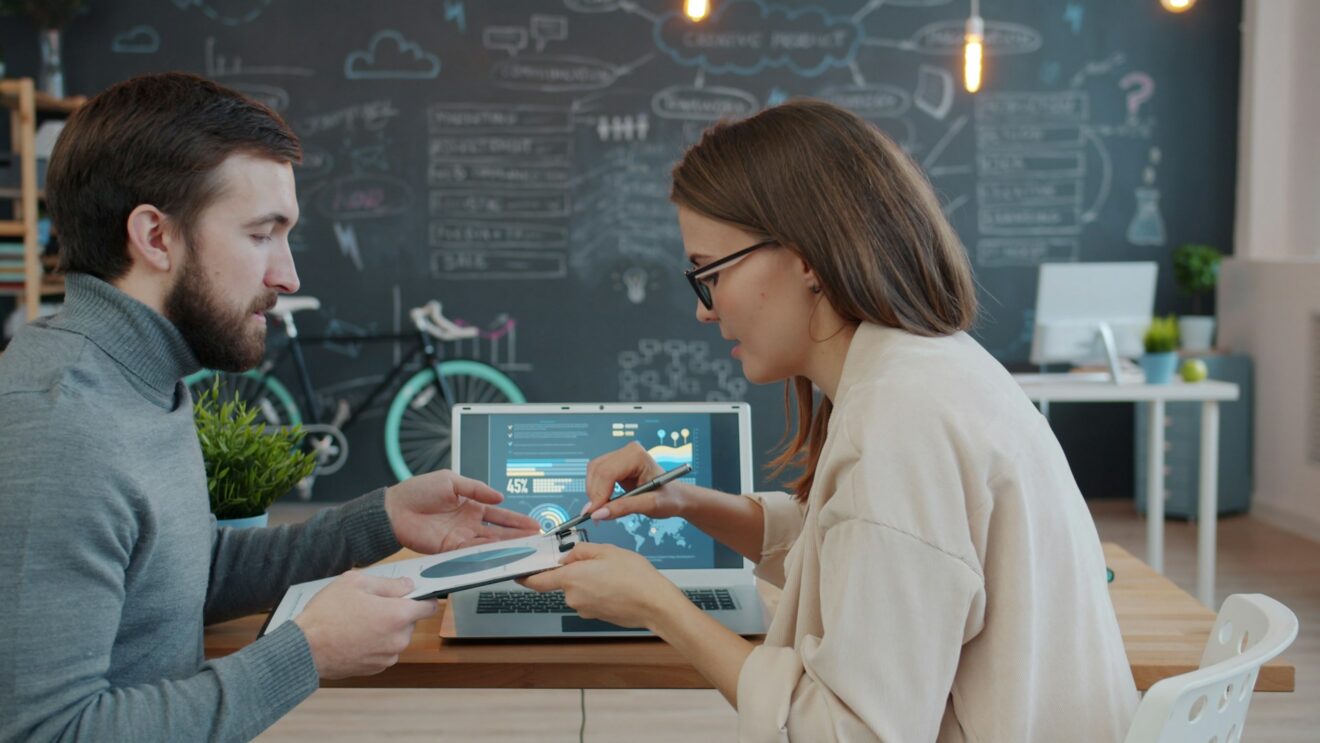 Male on left holding a tablet getting feedback from a female on the right. Both are seated. Used for a SmartBrief Marketing article on how companies can effectively build a marketing team.