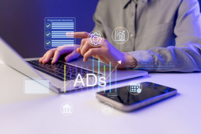 A person works on a laptop while focusing on programmatic advertising tasks. An electronic device lies nearby. Graphics appear on the screen showing data and stats related to advertising.