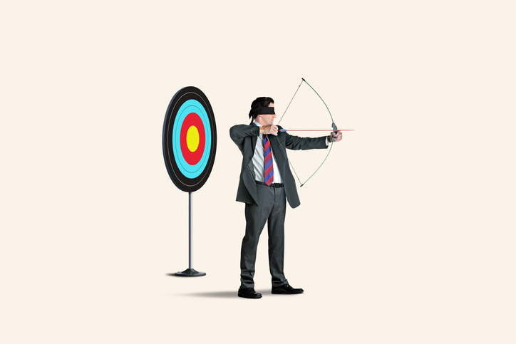 A blindfolded man with a bow and arrow and a target behind him aims in the wrong direction