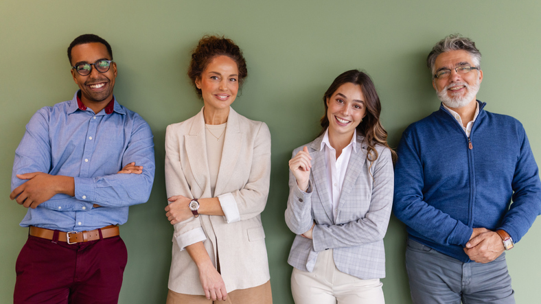 Diverse team of four professional colleagues of different ages and backgrounds looking at the camera in a modern office, representing teamwork, partnership, workplace diversity, and supportive corporate culture.
