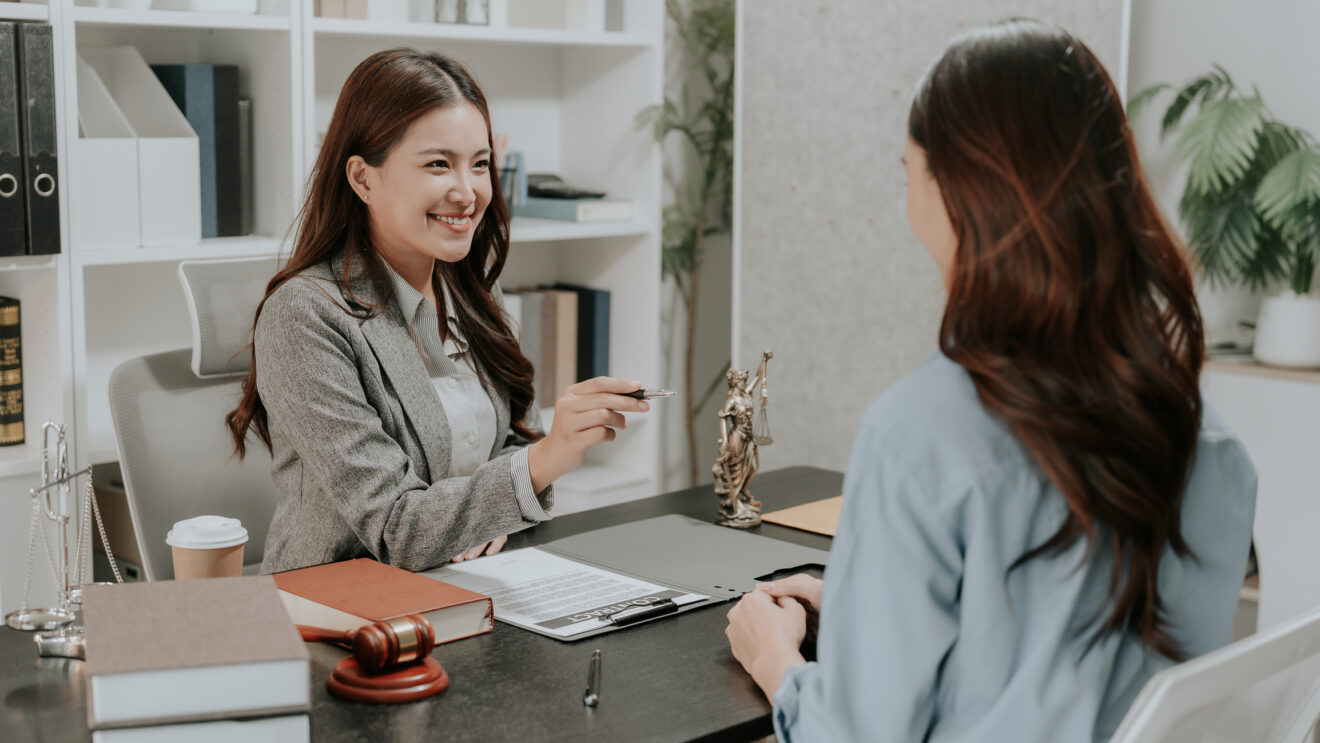 Professional female Asian lawyer holding law book and explaining legal contract details to a stressed client in corporate law firm office.